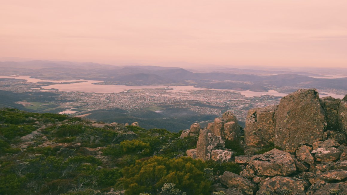 Photo of Mount Wellington’s view from the top with big rocky formations to the right and green trees to the left going downwards with a view of a city and a large body of water and small mountain ranges, with a pink hazy sky