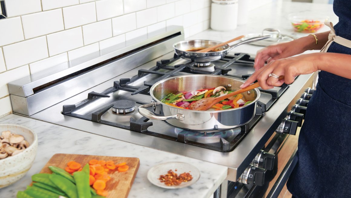 Photo of a person cooking mixed vegetables on a silver pot with a wooden spoon on top of a stove in a white-walled kitchen with a white marble countertop