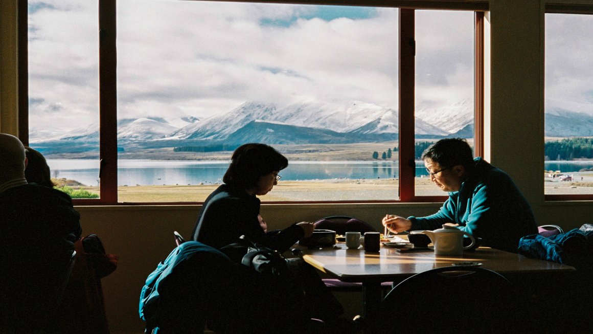 A middle-aged couple enjoying their New Zealand food by the large window showcasing Lake Tekapo, New Zealand and mountains in the distance and a sky covered with clouds and a blue sky barely visible behind the clouds