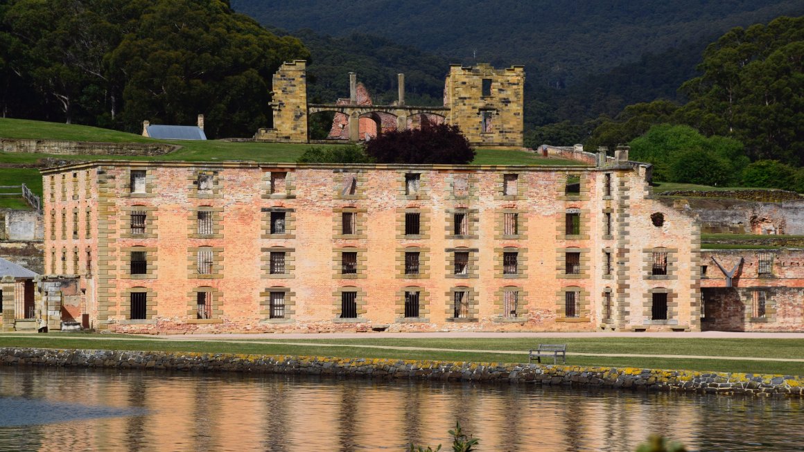 Photo of Port Arthur with an old shabby light orange-colored brick building with large barred windows and a flat top, with water in front of it and some grass in front and behind it, and in the background is an old run-down mostly destroyed yellow brick building, and in the distance are trees and mountains
