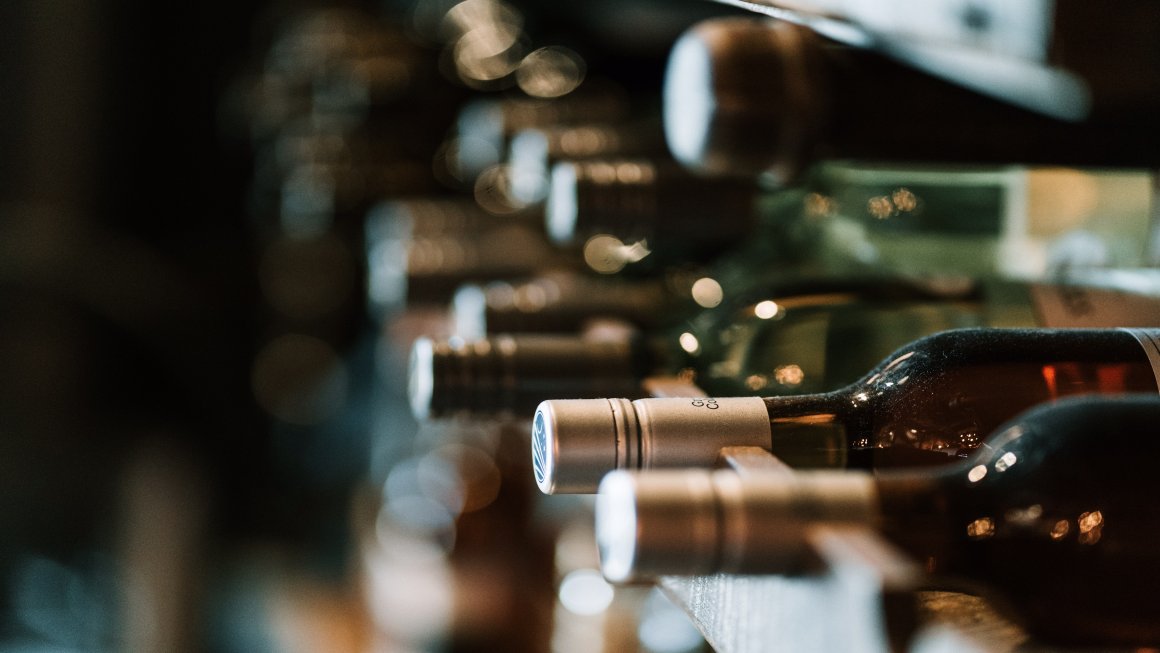 Closeup photo of wines in bottles and lying in rows in a cellar with differently colored bottles like maroon, dark purple, green, all of them with golden-colored caps, with two bottles nearest to the camera in focus and the rest blurred out with the background