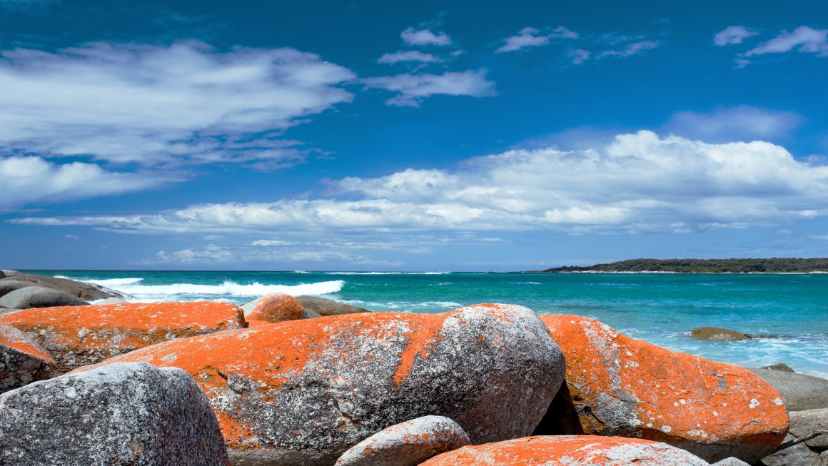 Photo of big rocks with bright orange tinges on top with a background of the blue ocean and a blue cloudy sky