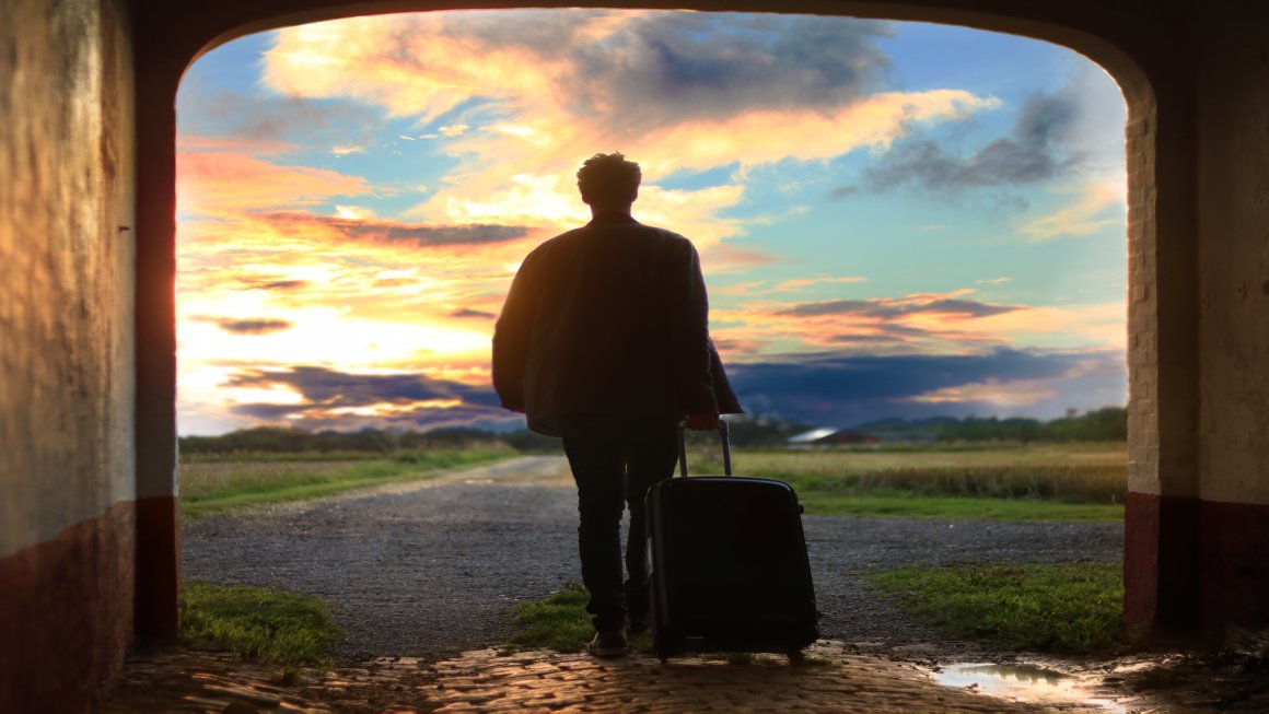 Photo of a man embarking on a trip wearing a jacket and jeans with a big stroller exiting from an archway towards an empty land with an empty road and grasslands and a blue and yellow warm sky with many clouds in the distance