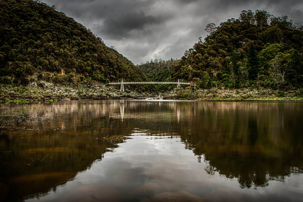 Prospect, Tasmania Lake Photo of a Green lake with mountains and forests in the distance and a big white bridge connecting the two mountains with the mountains and the land and bridge reflecting on the water, and a dark grey sky filled with clouds