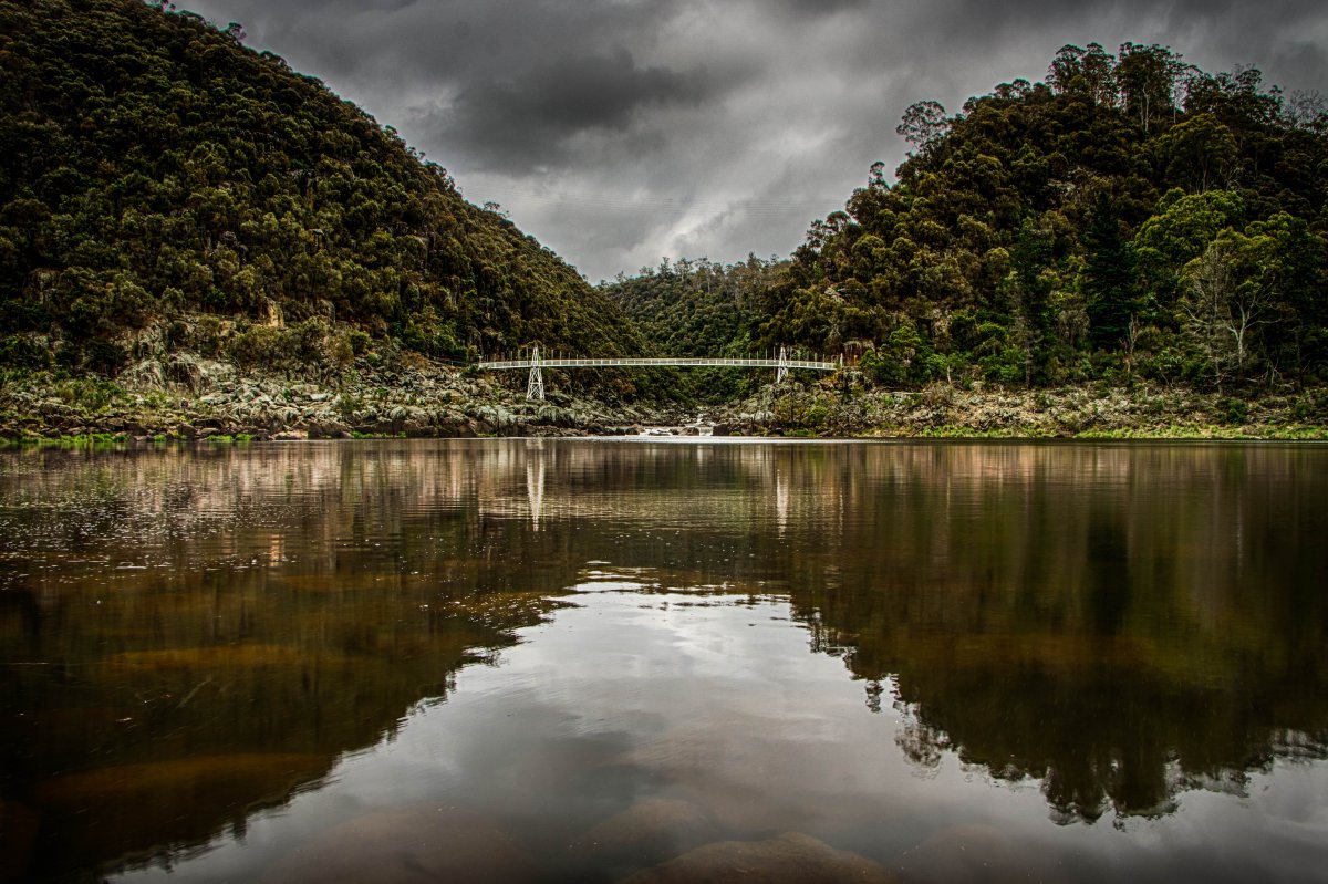 Photo of a Green lake with mountains and forests in the distance and a big white bridge connecting the two mountains with the mountains and the land and bridge reflecting on the water, and a dark grey sky filled with clouds