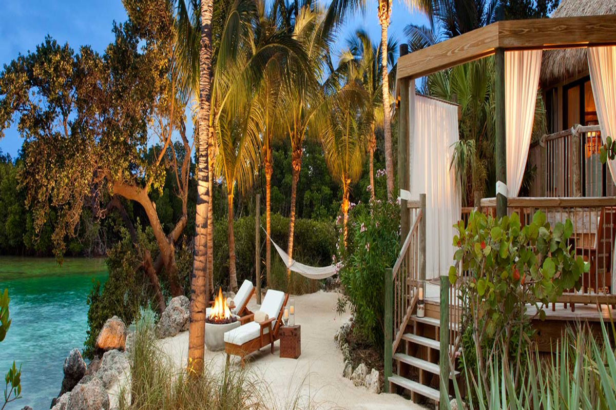 Two lounge chairs, a hammock, and a fire pit sit outside a bungalow amidst lush vegetation and water in Little Palm Island Resort & Spa, Little Torch Key, Florida